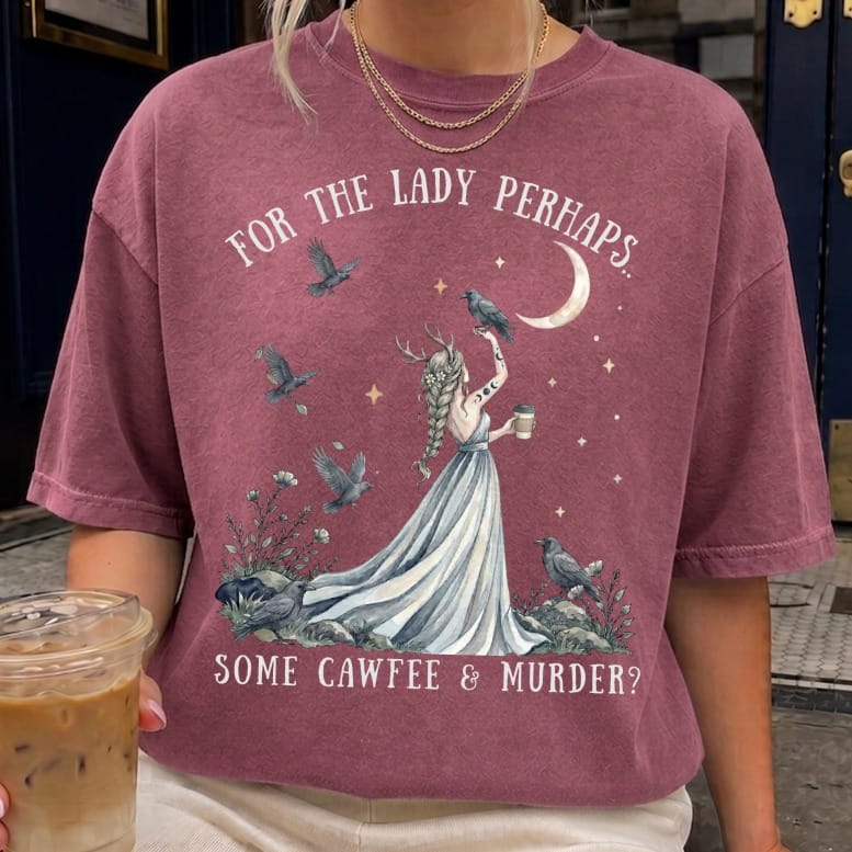 A person wears a mauve t-shirt featuring a graphic of a horned woman, crows, and text reading FOR THE LADY PERHAPS... SOME CAWFEE & MURDER?