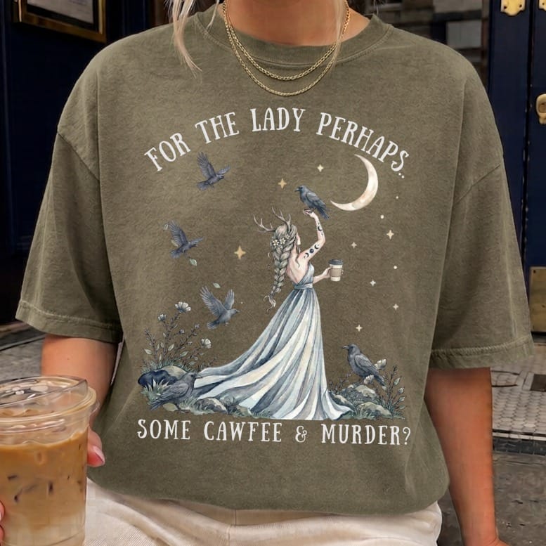 Olive green t-shirt featuring an illustration of a horned woman holding a coffee cup, surrounded by crows.