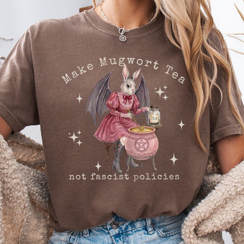Brown t-shirt with graphic of bunny in dress making mugwort tea, text reads "Make Mugwort Tea not fascist policies".