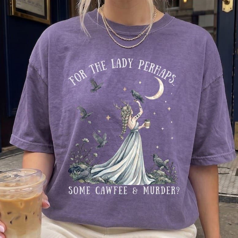 A person wears a purple t-shirt with a graphic of an antlered woman, crows, a crescent moon, and text reading For the lady perhaps, some cawfee and murder.