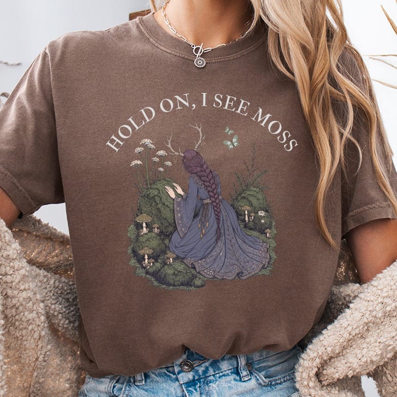 Brown t-shirt with "HOLD ON, I SEE MOSS" and forest illustration.