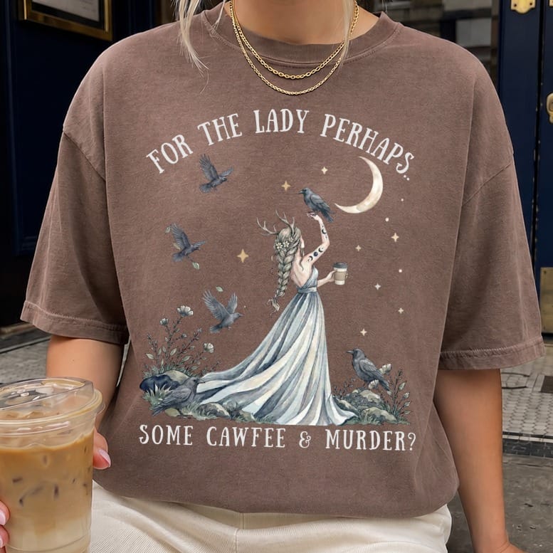 A person wearing a brown t-shirt featuring an illustration of a horned woman, crows, and text reading "FOR THE LADY PERHAPS... SOME CAWFEE & MURDER?".