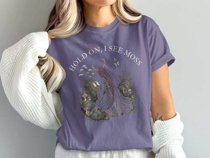 Purple t-shirt with a forest nymph illustration and text "HOLD ON, I SEE MOSS".